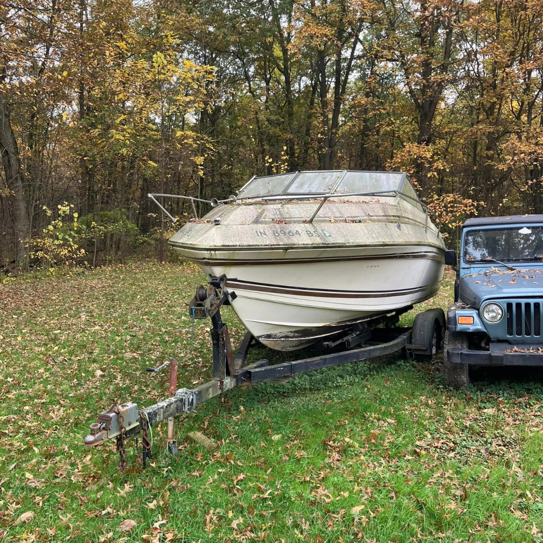 Junk boat sitting on a trailer next to a Jeep on a wooded Valparaiso property before removal by Patriot Clean Out