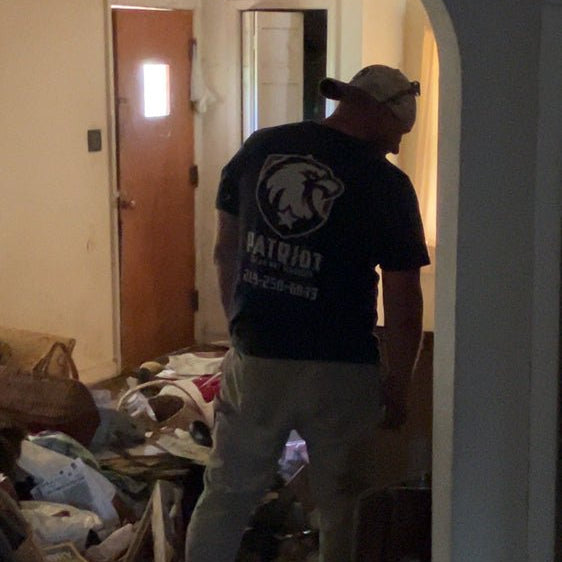 A Patriot Clean Out Services technician stands inside a cluttered home during a residential junk and hoarding cleanup in Northwest Indiana. The worker wears a company shirt with the Patriot logo while clearing debris, representing safe, efficient junk removal and home restoration services.